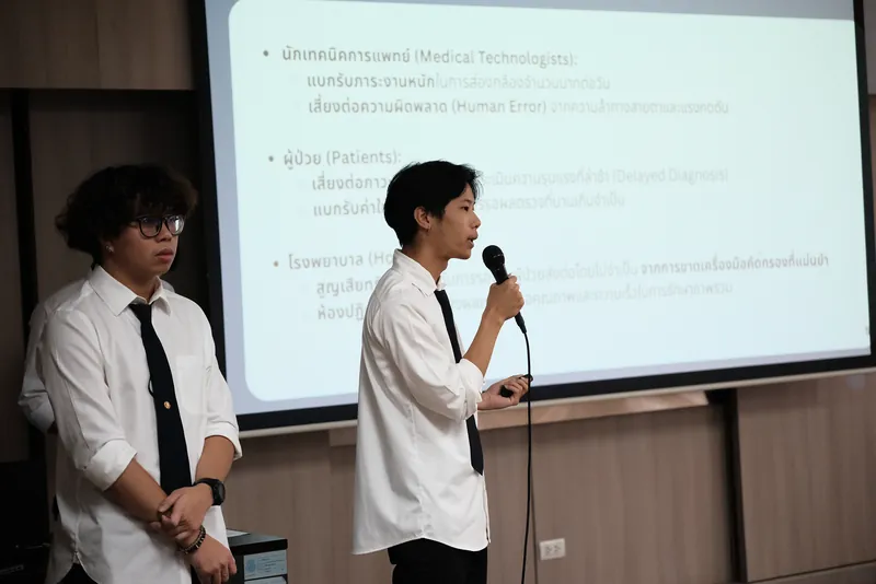 สมาชิกทีมสองคนยืนนำเสนอหน้าห้อง คนหนึ่งถือไมโครโฟนพูดอยู่หน้าจอฉาย